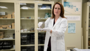 A woman in a lab coat (Elizabeth Benham) displays a silver-colored standard weight. 