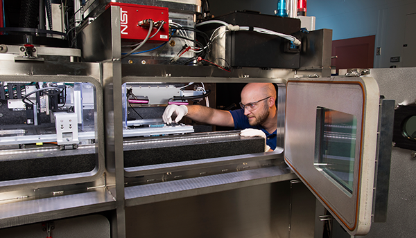 A man in safety glasses (Brandon Lane) reaches into equipment for metals-based additive manufacturing.