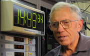 A man wearing glasses (Judah Levine) stands next to a digital time display.