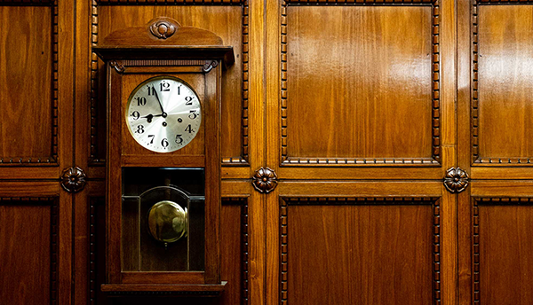 A wooden pendulum clock is mounted on wooden paneling. 