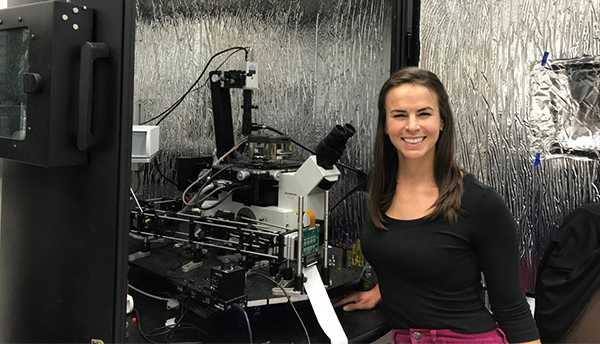 A woman (Callie Higgins) stands smiling in front of a piece of scientific equipment in a lab. 