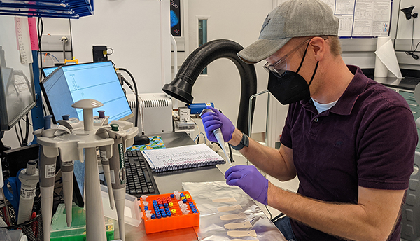 A man (Ed Sisco) in safety goggles and a baseball cap prepares samples at a laboratory bench.