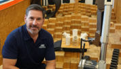 Adam Jacoff sits next to a metal robotic device with treads in a room with wooden blocks piled on the floor.