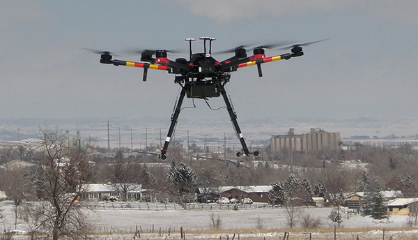 A black drone with red and yellow stripes hovers over a snowy landscape of houses and fields.