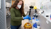 Woman in a green sweater and face mask works at a lab counter with an MRI phantom, a yellowish plastic sphere. 