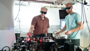 Two men wearing dark goggles (John Bollinger and Matt Affolter) stand at a table covered with scientific equipment. 