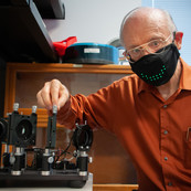 Paul Black, wearing a black mask, poses with a scientific device. 