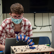 A woman wearing a face mask (Jennifer Case) bends over a laptop behind a wooden platform holding a flexible robot arm prototype. 