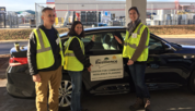 Three researchers in high-vis vests stand around a car with a sign on the door saying "Resilience."