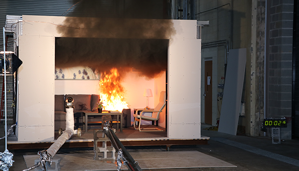 Black smoke pours out of a test room inside a larger facility, where an upholstered couch is in flames.