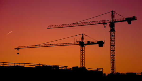 Two construction cranes are silhouetted against an orange sky.