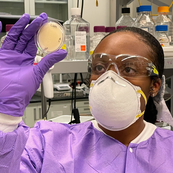 A scientist wearing protective gear examines a petri dish. 