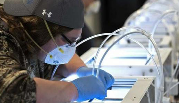 A person in a baseball cap and mask bends over a high-tech manufacturing workstation.