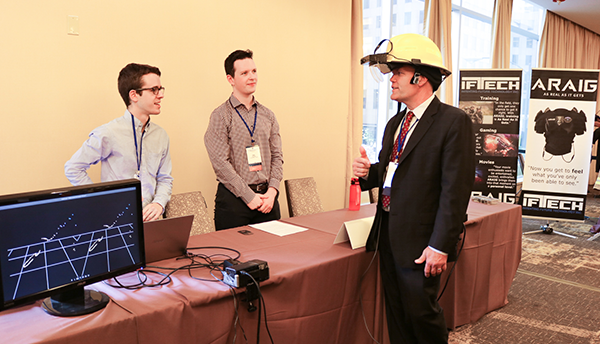 A man wearing a safety helmet (Dereck Orr) talks with two others standing behind a table at a PSCR conference. 
