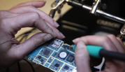 A close-up photograph shows a person's hands as they solder a wire onto a circuit board.