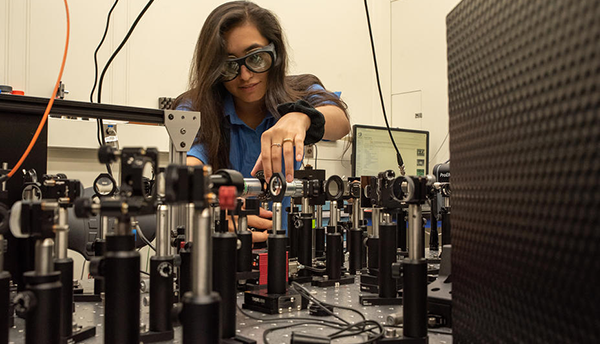 Valeria Viteri-Pflucker leans over a table full of optical equipment, wearing safety goggles.