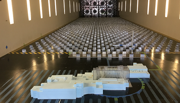 A model of a hospital building is positioned on the floor of a wind tunnel, a hallway structure with fans visible at the back.