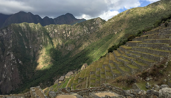 Sun and shade play over green and rocky mountains and Incan terraces outlined with stone walls. 