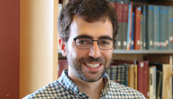 Adam Kaufman poses for a head shot with a shelf of library books in the background. 