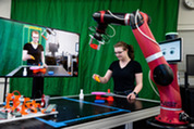 Megan Zimmerman moves plastic pieces on a table alongside a red robotic arm, while her image appears on a nearby screen.