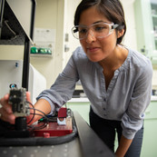 Polette Centellas wears safety glasses as she leans over a lab bench to manipulate a small device attached there.