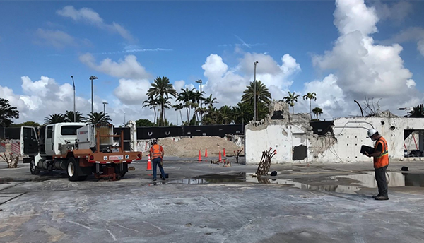 A flatbed truck with "thumper" equipment on the back sits in a puddled parking lot near the remains of Champlain Towers South.
