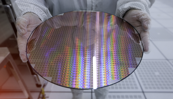 A person wearing a white coverall and gloves holds out a circular, reflective wafer of silicon. 