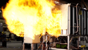 A ball of flame bursts out of a metal box on legs in a lab, with a digital readout and a tripod placed underneath. 