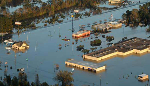 The tops of houses and larger buildings are visible above floodwaters in an aerial view of a flooded landscape.