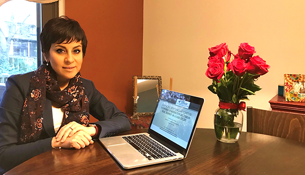 Yalda Saadat poses at home, sitting at a table with an open laptop and a vase of flowers. 