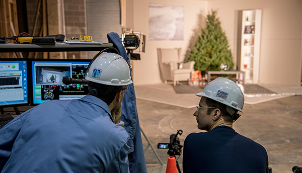 Two people wearing hard hats sit behind computers while looking over at a video set of a living room with a Christmas tree.