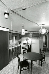 B&W vintage photo shows a man in office attire looking at a scientific device on the counter of a kitchen in a mobile home. 