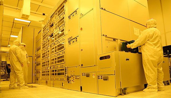 Three people in white coveralls stand at a large, floor-to-ceiling machine with panels and glass windows in a room with yellow lighting.