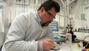 John Kitching leans over a table in the lab, using tweezers to point to part of a chip-scale device in a clear plastic box. 