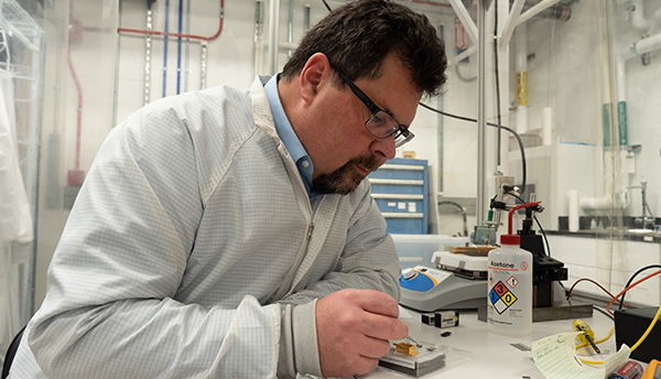 John Kitching
                                          leans over a table in the lab,
                                          using tweezers to point to
                                          part of a chip-scale device in
                                          a clear plastic box. 