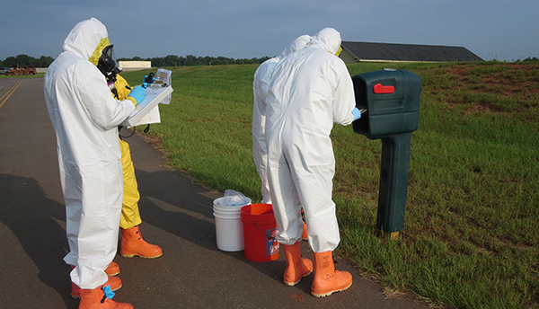 Four figures in coveralls stand outdoors around a mailbox, looking at a clipboard and other items, during a biohazard training exercise.