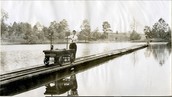 Historical photo shows a railway stretching over the surface of a small lake, with a small railcar being moved by a man in a tie.
