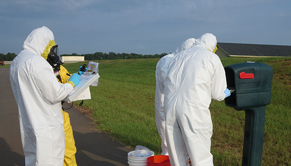 Four figures in coveralls stand outdoors around a mailbox, looking at a clipboard and other items, during a biohazard training.