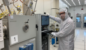 A NanoFab intern in a white coverall and safety glasses poses standing next to a rack of computers and other devices in a lab.