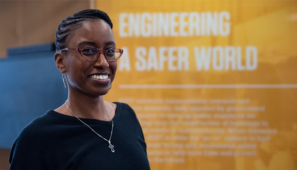 Jazalyn Dukes poses smiling in front of a poster that reads "Engineering a Safer World."