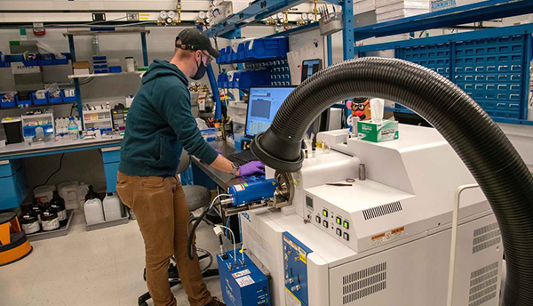 Ed Sisco in the lab stands at a computer alongside a large scientific instrument with a curving black tube in the foreground.