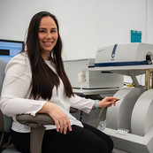 Ileana Pazos poses sitting next to a large gray machine and holding a small device pointed toward two cylindrical parts of the machine.