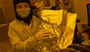 Jessie Zhang, wearing safety glasses and coveralls, holds a piece of aluminum foil in the yellow light of a clean room. 