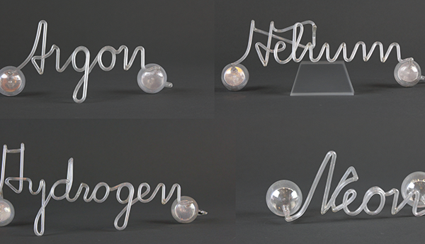 Glass tubing is shaped to spell the names of four noble gases in cursive: Argon, Helium, Hydrogen, Neon 