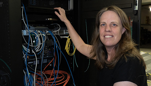 Jeanne Quimby crouches next to a rack of computer equipment with many different colored cord connections. 