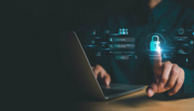 A person sitting with a laptop reaches out to touch a padlock icon floating in the air with other cybersecurity symbols.