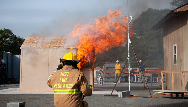 A NIST firefighter in safety gear watches a shed burn next to a structure representing a wall of a residence during an experiment.