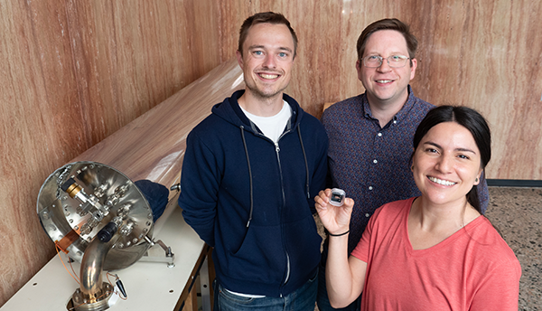 A woman stands with two men, holding up a small device, a chip scale atomic clock. To their right is a large cylindrical metal instrument.