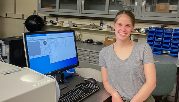 SURF student Grace Waters poses smiling in the lab, leaning against a desk with a computer monitor showing data and graphs. 