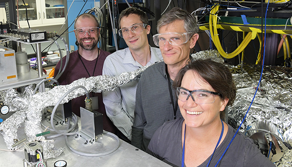 Four CAVS researchers pose standing in a lab, smiling and wearing safety glasses, surrounded by wires and other electronics.